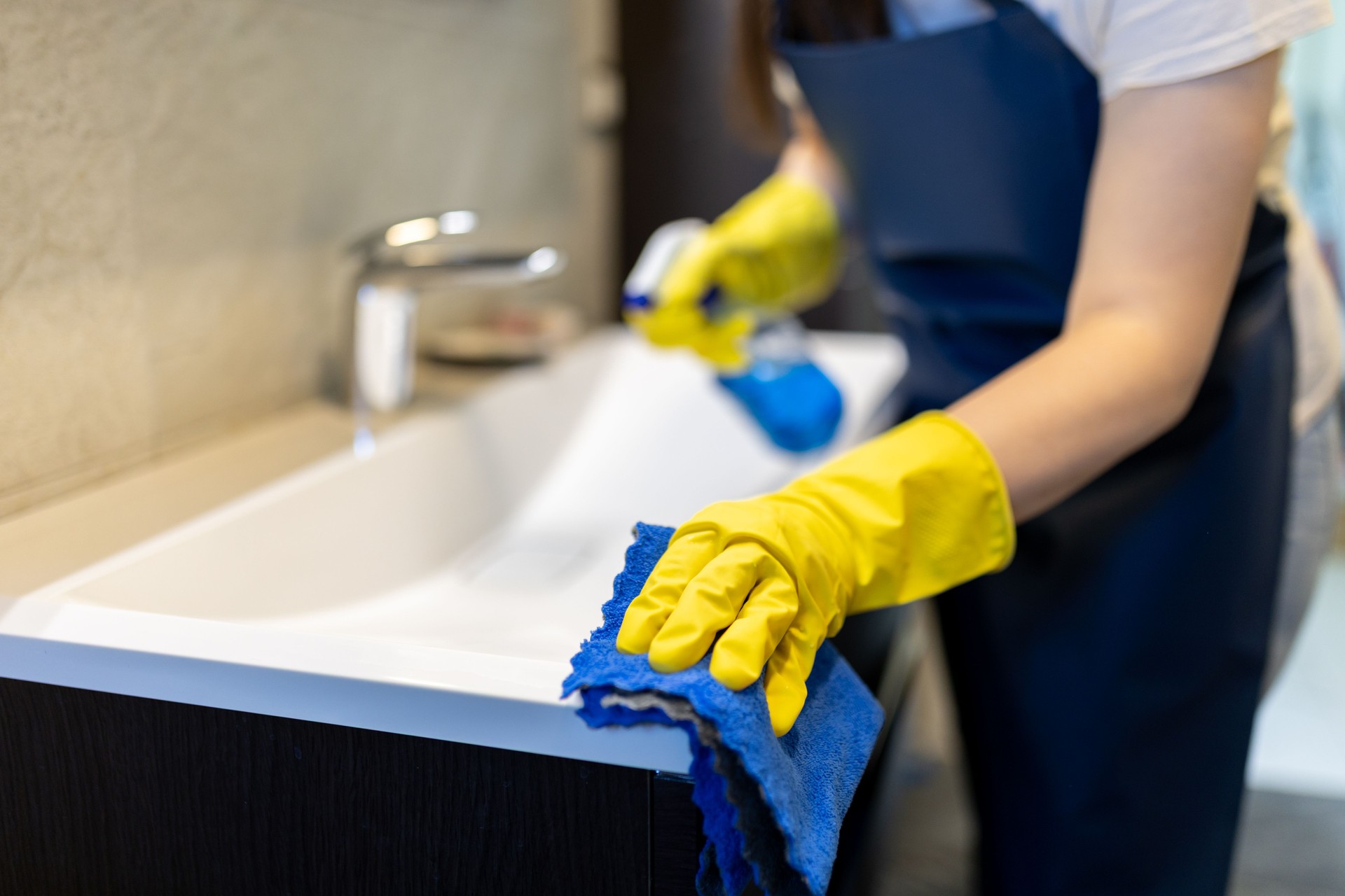 Cleaning a bathroom sink with gloves and a cloth in a bright, modern space