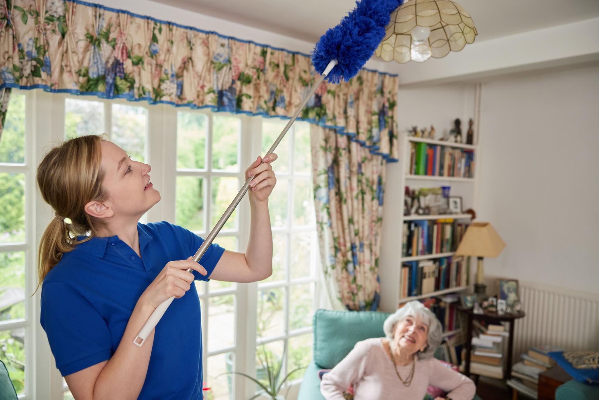 Female Home Help Cleaning And Dusting House Talking To Senior Woman